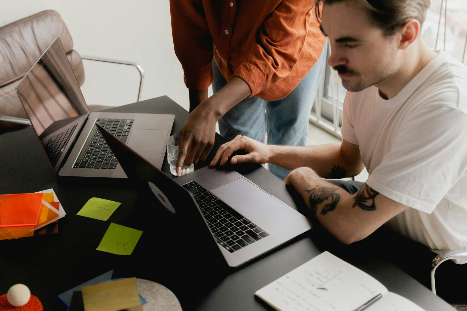 A high-angle view of two people working together at a dark office table. A seated man with a mustache and tattooed arms looks closely at his open laptop, while a standing colleague in an orange button-down shirt points at his screen. The workspace features a second open laptop, green sticky notes, orange color swatches, a glass of water, and an open notebook with handwritten notes and diagrams.