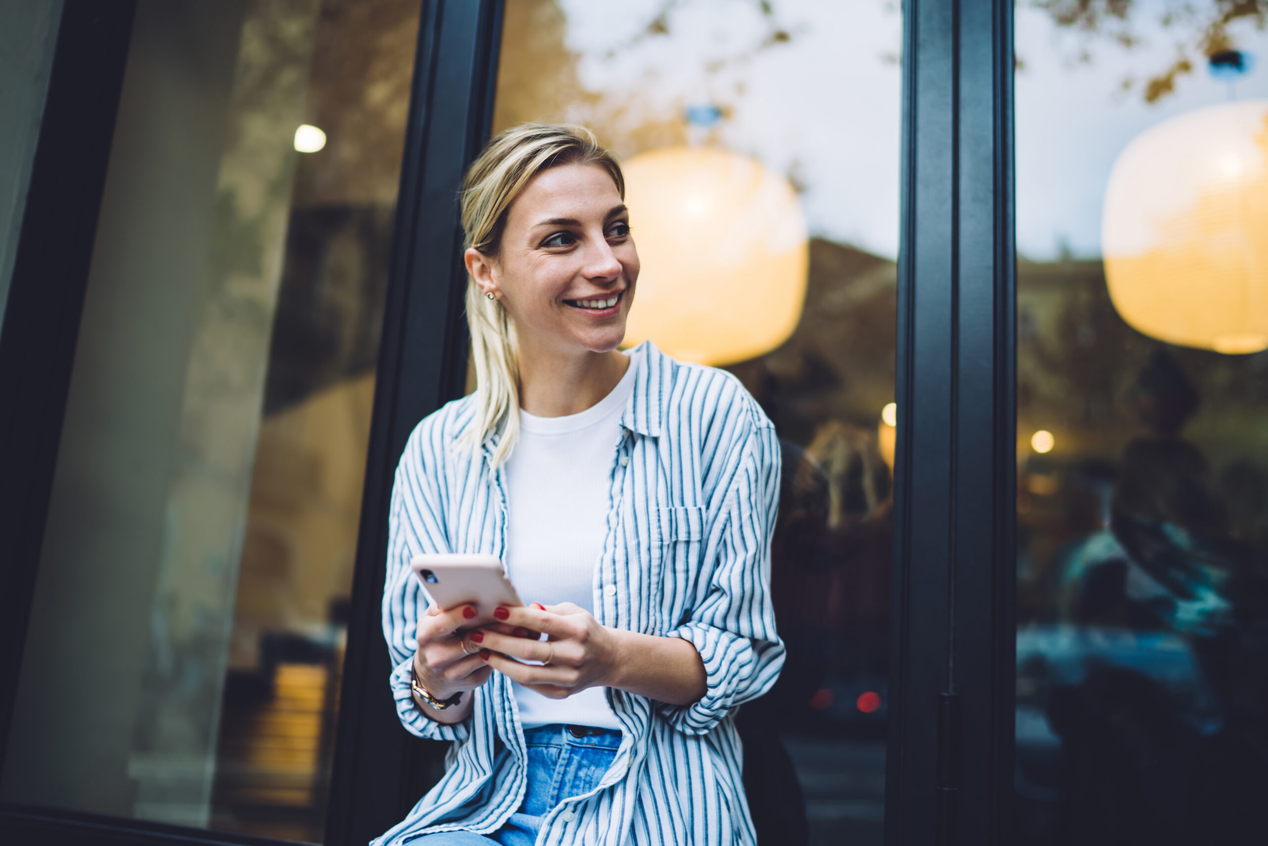 A smiling young woman holding a smartphone outdoors, checking social media apps outside a cafe storefront.